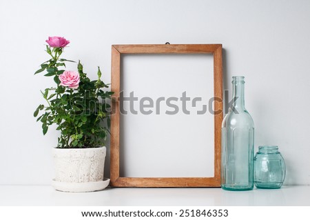 Vintage blank wooden frame, bottles and rose in a pot on a white wall