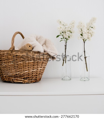 Wicker basket with home textiles and white lilac on a white chest, white vintage room interior decoration.