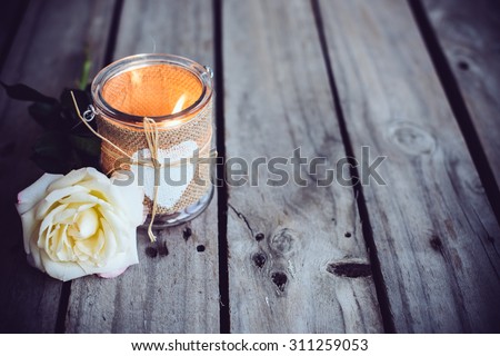 Burning candle in a decorative jar and flower of rose on old wooden board. Rustic decor.