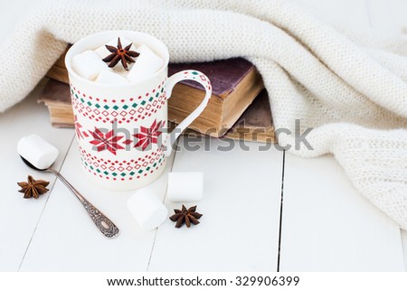 Cozy winter home background, cup of hot cocoa with marshmallow and star anise, old vintage books and warm knitted sweater on white painted wooden board background.