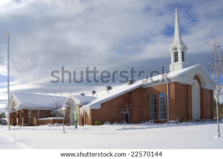 A red brick church covered in snow, standing in a snowy field with its steeple against a blue break in the cloudy sky.