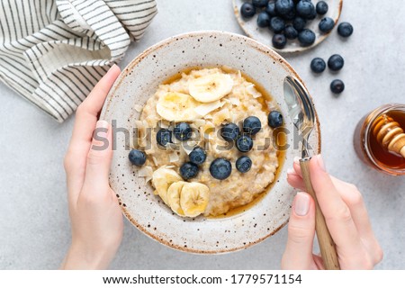 Oatmeal porridge with banana, blueberries and honey. Female hands holding bowl of oatmeal porridge over grey concrete background, table top view Photo stock © 