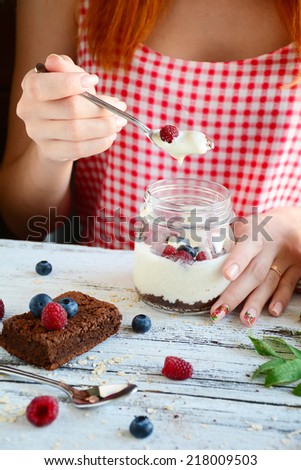 Redhead girl in red morning dress eating creamy dessert in jar with brownies and yogurt topped with fresh raspberries and blueberries,