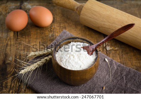 White wheat flour in brown wooden bowl, fresh eggs, wheat ears and rolling pin on wooden table. Cooking at countryside, food ingredients