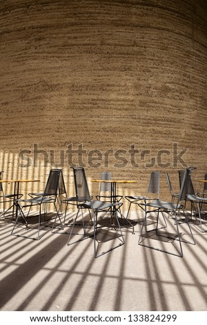 Wide angle view of metal Chairs and tables and clay wall, lit by natural light through gaps between bars, as suggested by the shadows.