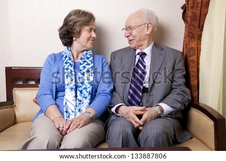 A high society senior couple (he\'s in his 80\'s, she\'s in her late 60\'s) sitting on a sofa looking at each other with very much love and joy.