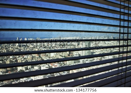 Defocused venetian blinds in their open position, revealing a dirty window with Tel Aviv buildings and the Mediterranean sea in the background.