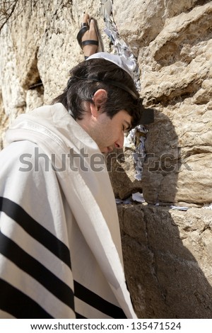 A Jewish adult man wearing a Jewish praying shawl, Yarmulke and Phylacteries on his head and left hand; praying in front of the  holy Western Wall (Wailing Wall) in the old city of Jerusalem, Israel.