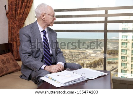 An elderly (in his 80\'s) business man wearing suit and tie sitting in a hotel\'s business lounge, looking through the window for a little break from the documents he is inspecting.