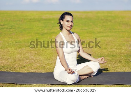 An adult woman sitting on a black yoga mattress in the Siddhasana (aka Accomplished) pose, on a green lawn with cloudy blue sky in the background.