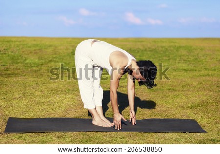 An adult woman standing on a black yoga mattress in the Uttanasana (aka standing forward bend) pose, on a green lawn with cloudy blue sky in the background.