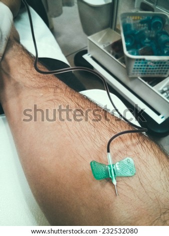 1st person POV view of a right arm with a needle stuck in it; taking blood for a blood test. In the background medical equipment can be seen, indicating it is in the nurse\'s room.