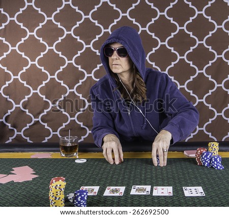 A woman sitting at a poker table wearing sunglasses and hoodie playing cards with a brown background