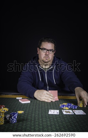A man sitting at a poker table wearing a hoodie gambling playing cards against a black background
