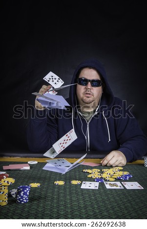 A man sitting at a poker table wearing a hoodie gambling and throwing his cards against a black background