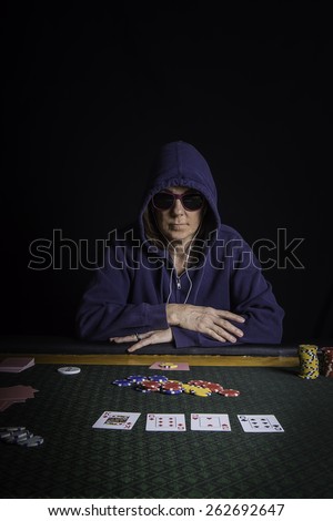 A woman sitting at a poker table wearing sunglasses and hoodie playing cards with a black background