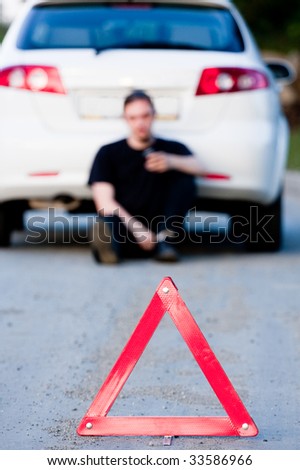 Young man sends an sms sitting by a white car. Focus is on the red triangle sign