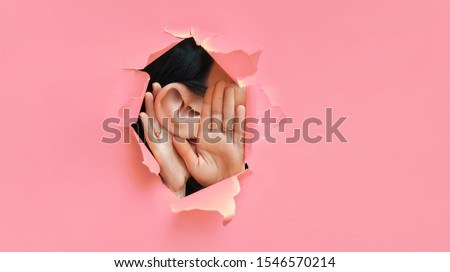 Female ear and hands close-up. Copy space. Torn paper, pink background. The concept of eavesdropping, espionage, gossip and the yellow press. Caricature with an enlarged ear.