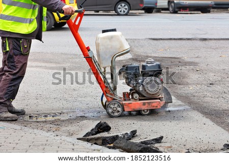 A road worker in reflective overalls uses a portable asphalt cutter to cut worn asphalt with a diamond blade to repair part of the roadway. Foto stock © 