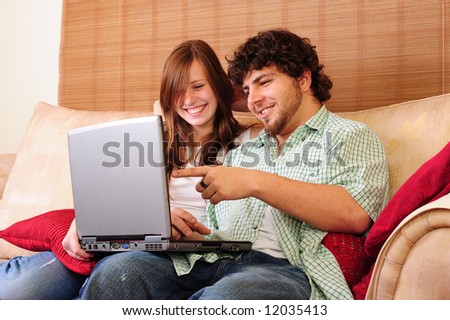 Young couple relaxing on a couch, playing with a laptop computer, laughing and pointing.