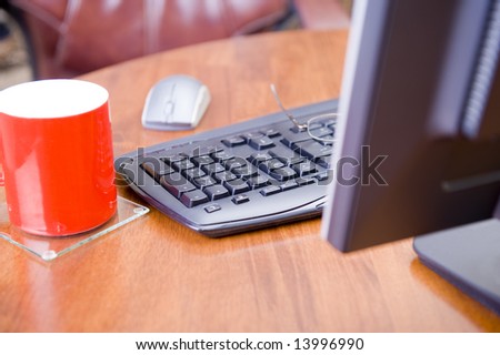 A keyboard, mouse and monitor standing on an executive wooden desk, with a cup of coffee on the side on a glass coaster.