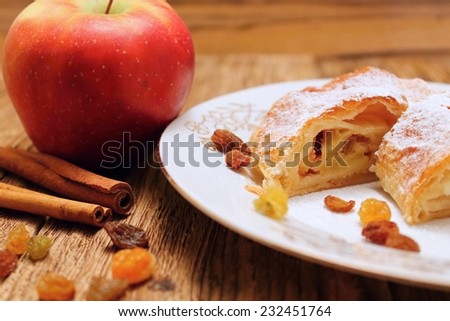 Apple pie strudel on white plate placed on old worn wooden table with apples and cinnamon and with raisins around