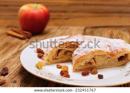 Apple pie strudel on white plate placed on old worn wooden table with apples and cinnamon and with raisins around
