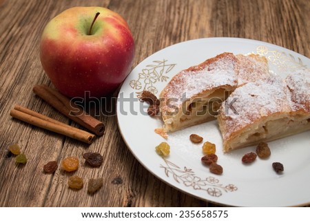 Photo of two portions of apple pie strudel on white plate with golden motif on old wooden board with cinnamon and raisins around and with one red apple near the plate.