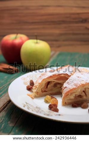Two pieces of apple pie strudel on white plate with raisins around and with two apples and cinnamon in back placed on green worn wooden table.