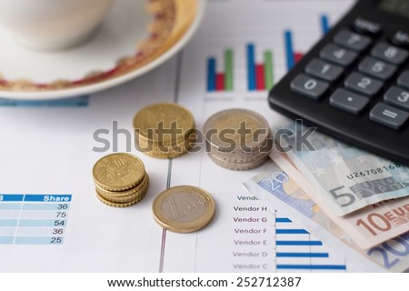 Horizontal photo of euro coins in stacks and bills under calculator with coffee cup in background placed on paper sheets with bar charts.