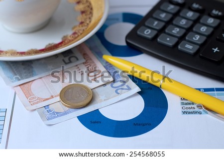 Horizontal photo with paper sheet with printed pie chart, euro bills under coffee cup, yellow pen, black calculator and one euro coin. Photo is with blue tone.
