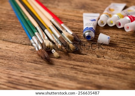 Horizontal photo of Opened blue acrylic color and set of paintbrushes with other colors in tubes placed on old worn wooden board