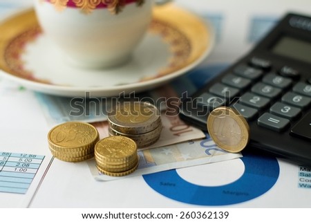 Horizontal photo of Three stacks of coins on other bills placed on paper sheets with pie chart with black calculator and tea cup.