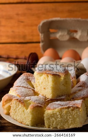 Vertical photo of plate full of curd cake portions on white plate with bowl full of powder sugar and eggs with paper box in background.