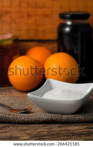 Vertical photo of Bowl with white yogurt on jute cloth and spoon on left, three oranges and bottle of marmalade on old worn wooden table.