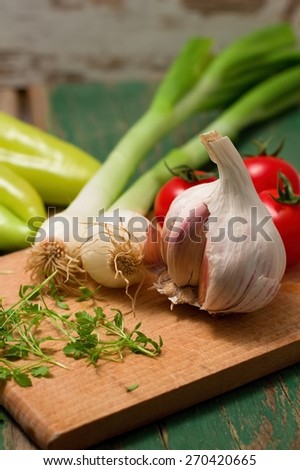Vertical photo of Garlic, spring onion and cress on wooden board which is placed with other vegetable - tomatoes and paprica on green table