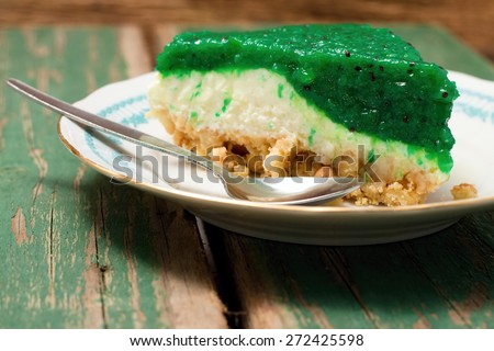 Horizontal photo of single portion of green fruit kiwi cake with visible seeds in jelly layer. Cake is placed on white plate together with spoon and everything is on old wooden table with worn color.