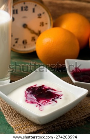 Vertical photo of Table with prepared breakfast with white yogurt in square bowl plus berry fruit inside. Glass of milk and oranges are in background with alarm clock.