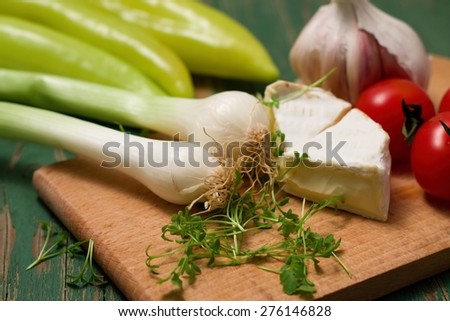 Horizontal photo of two heads of spring onion. Cress herb, two portions of camembert, tomatoes, garlic and peppers are around. All is placed on chopping board and green wooden table.