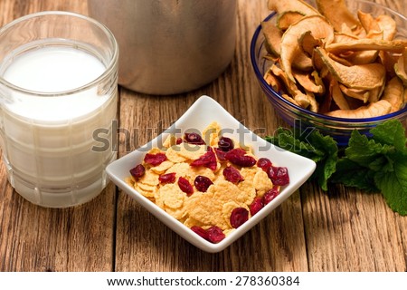 Horizontal photo of modern square bowl full of cornflakes with fruit. Glass of milk and other bowl with dried apple are around. All is placed on old wooden table.