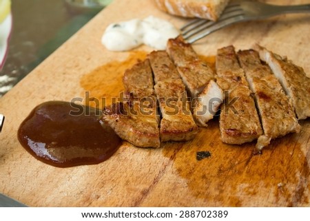 Horizontal photo of pork steak cut on stripes with bbq sauce in front and tatar sauce in beck. Silver fork is in background and all is placed on wooden chopping board.