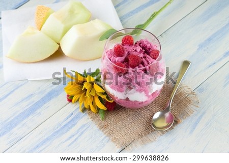 Horizontal photo of glass with two kinds of ice cream - vanilla and fruit on jute burlap cloth. Yellow flower, spoon and galia melon are around on light blue wooden board.