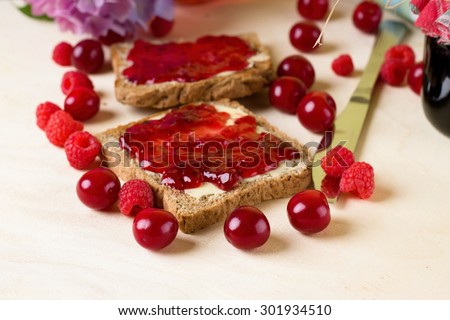 Horizontal photo with couple of toasts with butter and fruit homemade marmalade from raspberries and cherries which are spilled around on light blue wooden board.
