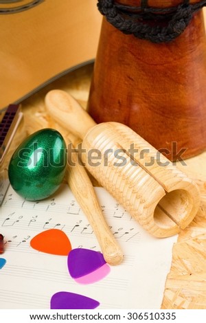 Vertical photo of wooden guiro and green egg shaker on piece of paper with written music. Bongo, acoustic guitar and few picks are around on OSB board.