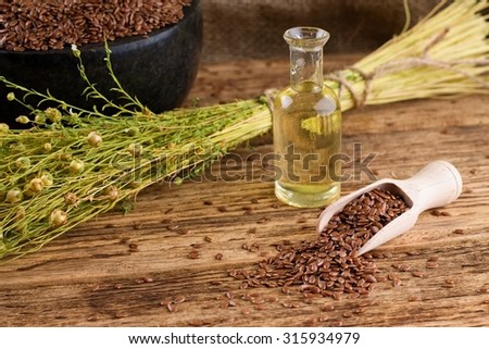 Horizontal photo with wooden spoon full of flax seeds next to small glass bottle with flax oil and bunch of flax plants. In background is marble bowl with seeds and piece of burlap.
