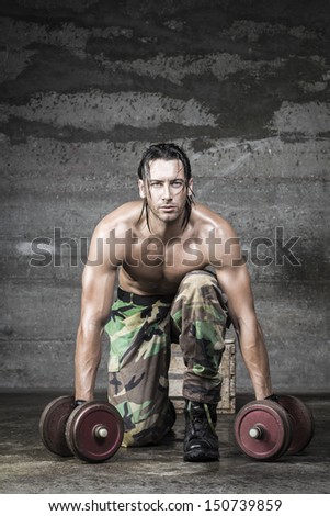 portrait of muscle athlete with camouflage pants looking at camera and weightlifting on wall background