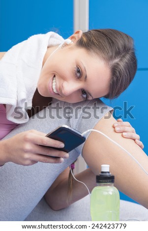 Fitness smiling young woman with towel and bottle using her smart phone with earphones in blue dressing room