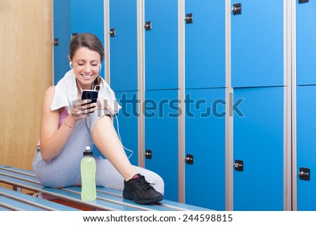 Fitness young smiling woman with towel and bottle using her smart phone with earphones in blue dressing room