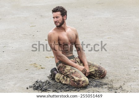 Muscled Shirtless Soldier in Camouflage Pants and Black Shoes Kneeling on the Beach Sand