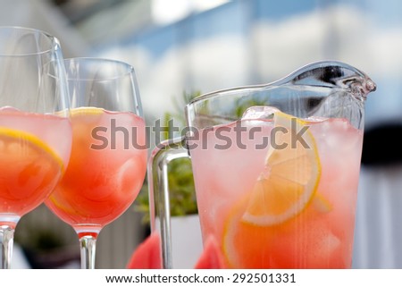 Watermelon juice: cold pink drink in glass jug. wine glasses and pitcher with sweet Watermelon lemonade and citrus slices, two glasses. Sun lights. Outdoor, summer party, day. Side view. Soft focus.